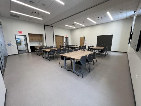 A room containing 6 pods of tables with seating for 36, 2 television monitors, a sink, and a minifridge. 